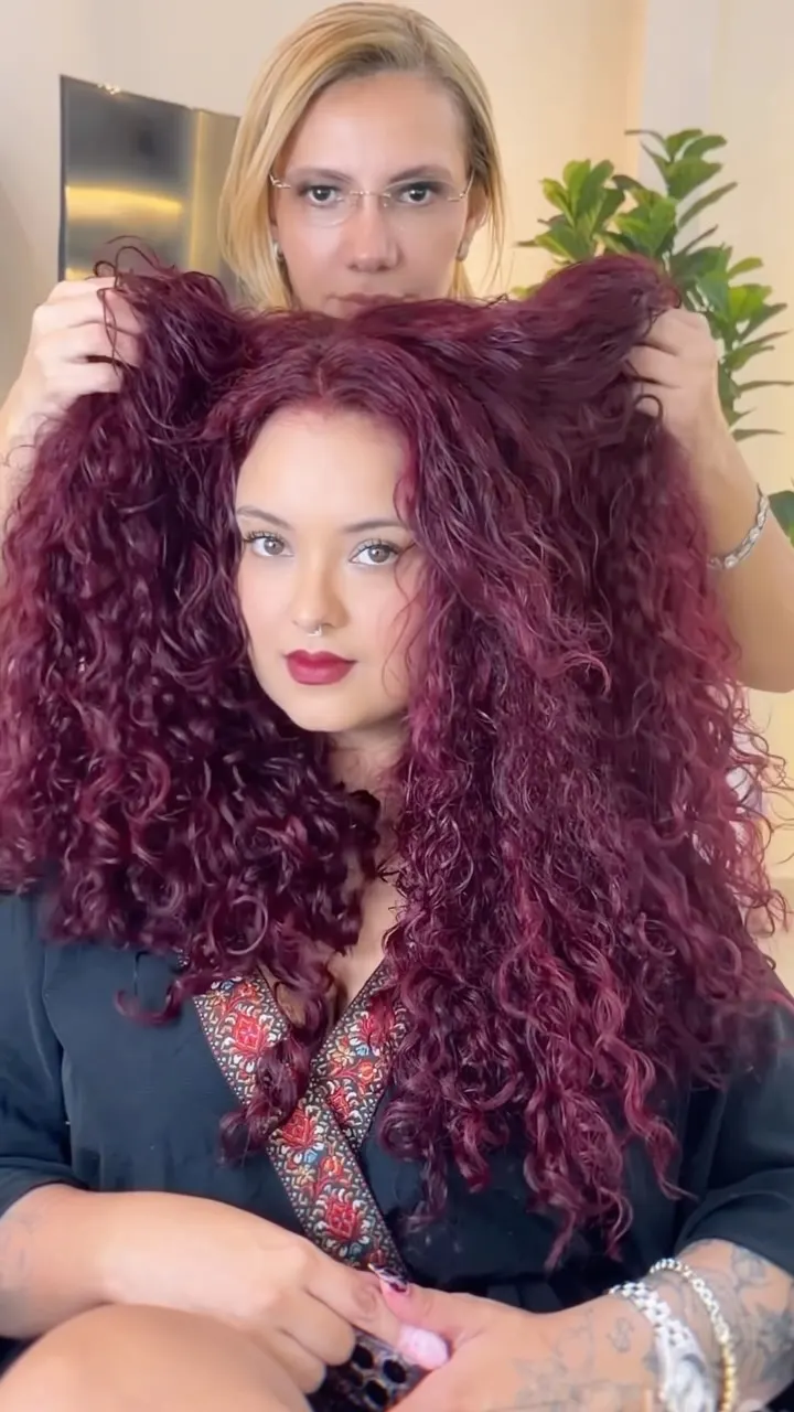 Cliente com cabelo cacheado volumoso em tom vinho, com a profissional ao fundo.