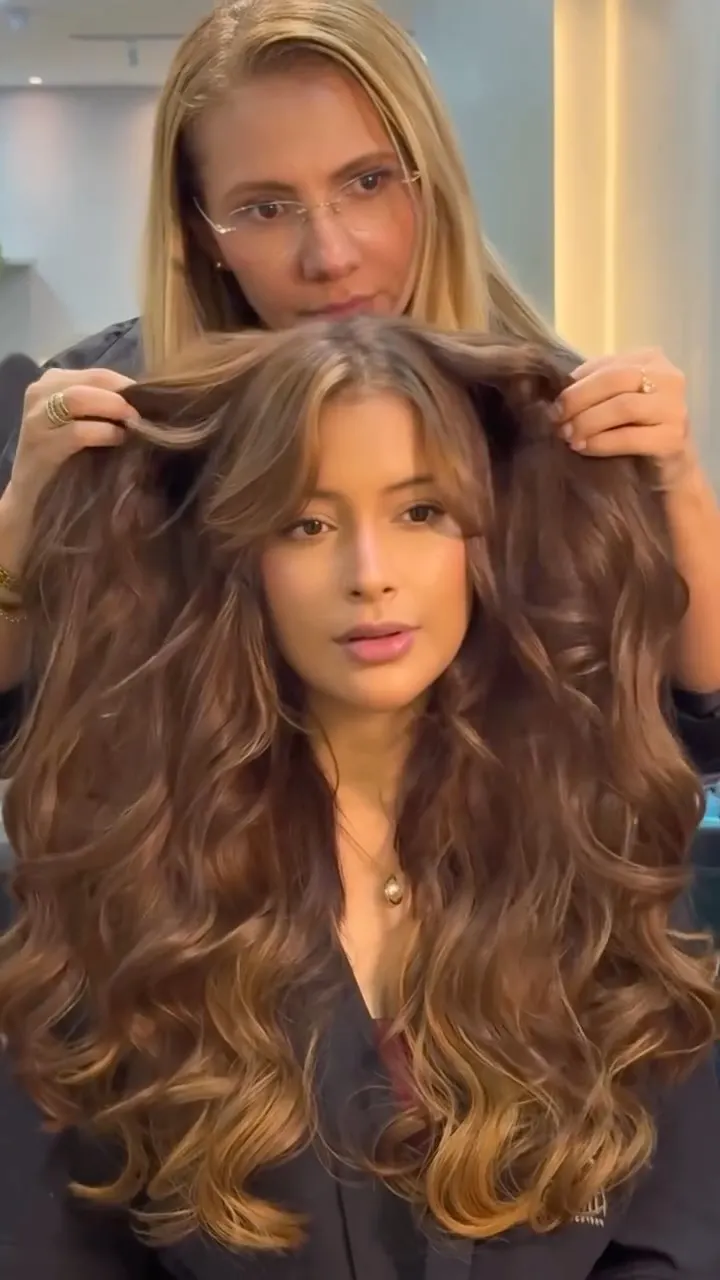 Cliente com cabelo castanho mocha longo e ondulado, com a profissional ao fundo.